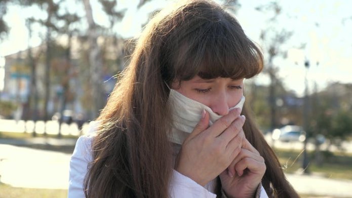 A woman pulling down her face covering to cough into her hand.