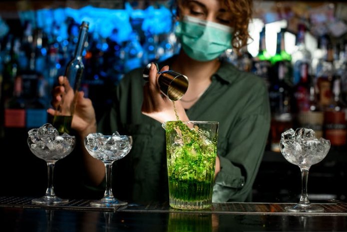 A bartender pours a cocktail while wearing a face mask.