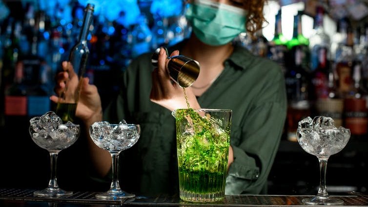 A bartender pours a cocktail while wearing a face mask.