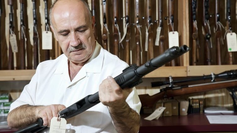 A man cocks a rifle, standing in front of a rack of rifles in a US gun shop.