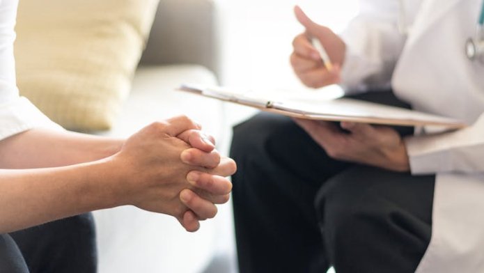 A person with clasped hands sits opposite another with a clipboard.