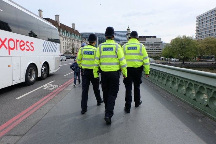 View from behind of three Met police officers walking side by side across a bridge, all wearing high-vis Met police jackets