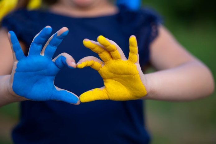 A young girl making the shape of a heart with her hands, one painted blue and one painted yellow like the Ukraine flag