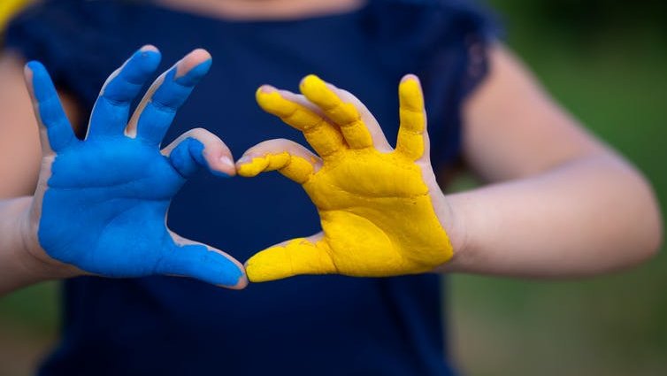 A young girl making the shape of a heart with her hands, one painted blue and one painted yellow like the Ukraine flag