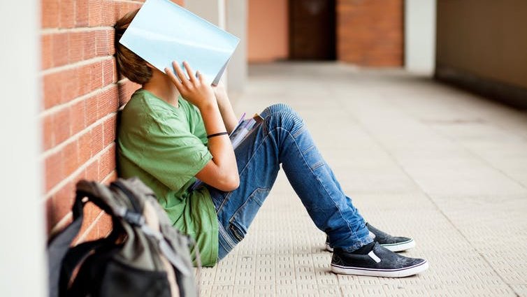 A teenage boy in a green t-shirt and jeans sits on the ground holding a book over his head.
