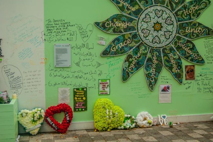 A green-painted section of wall with a floral mosaic, floral tributes and written messages.