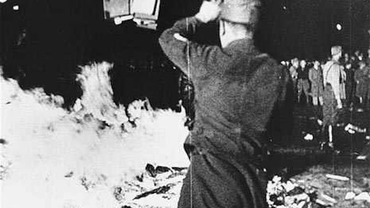 Black and white photo, a Nazi official viewed from behind throws books onto a fire.