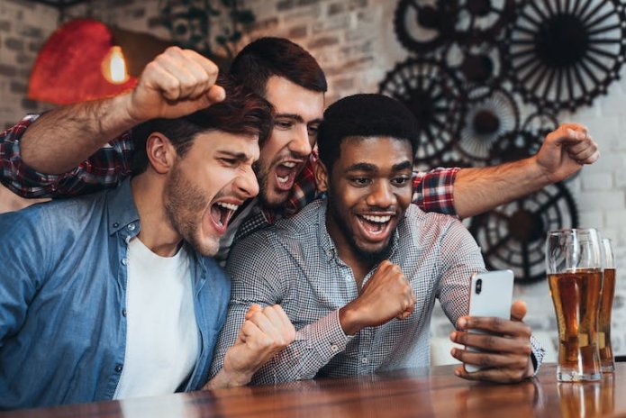 Three men cheer while gathered around a mobile phone, they are sitting at a bar with a pint of beer