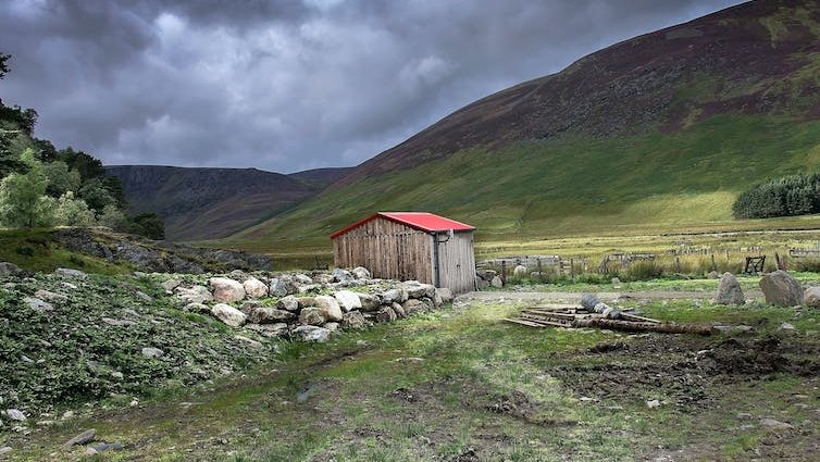 A rural hut with a red roof in mountainous countryside under a cloudy sky.