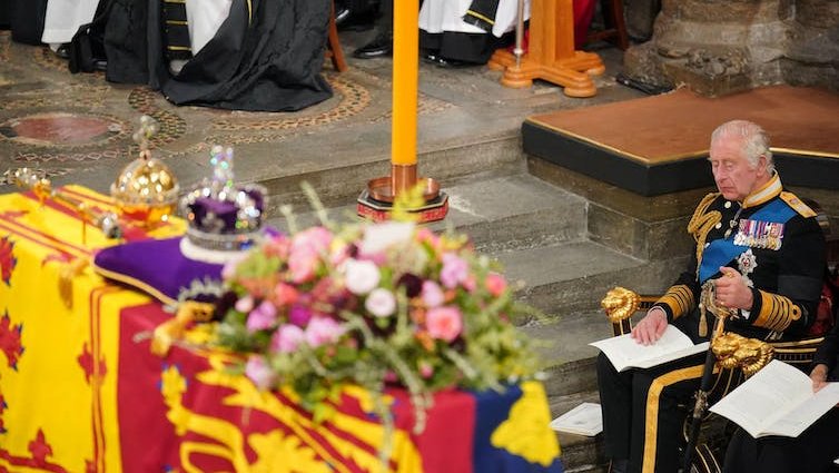 A coffin draped in the royal standard with a purple cushion, a candle and a crown, and the king seated beside it.