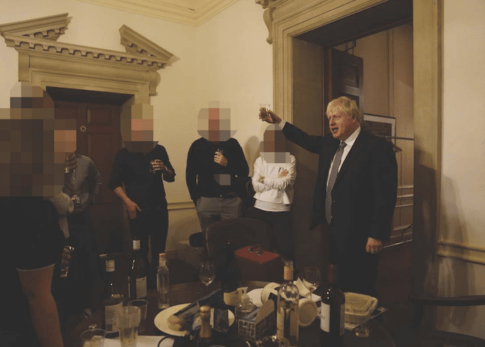 Boris Johnson raising a glass next to colleagues in front of a table covered in wine bottles.