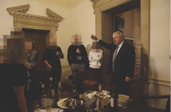 Boris Johnson raising a glass next to colleagues in front of a table covered in wine bottles.
